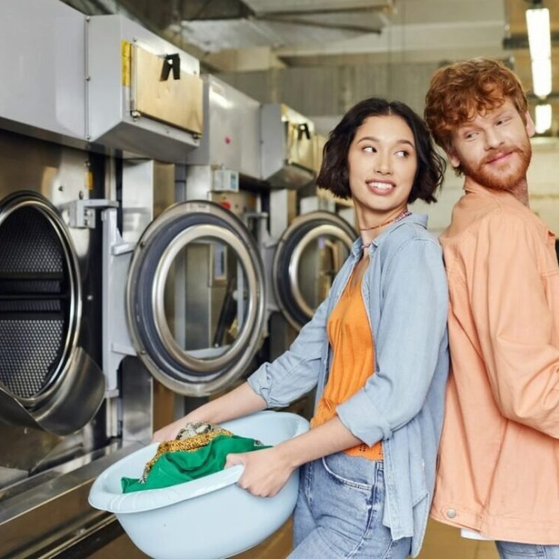 imgi_65_smiling-multiethnic-couple-standing-back-to-back-and-holding-basins-with-clothes-in-coin-laundry-e1716535733496